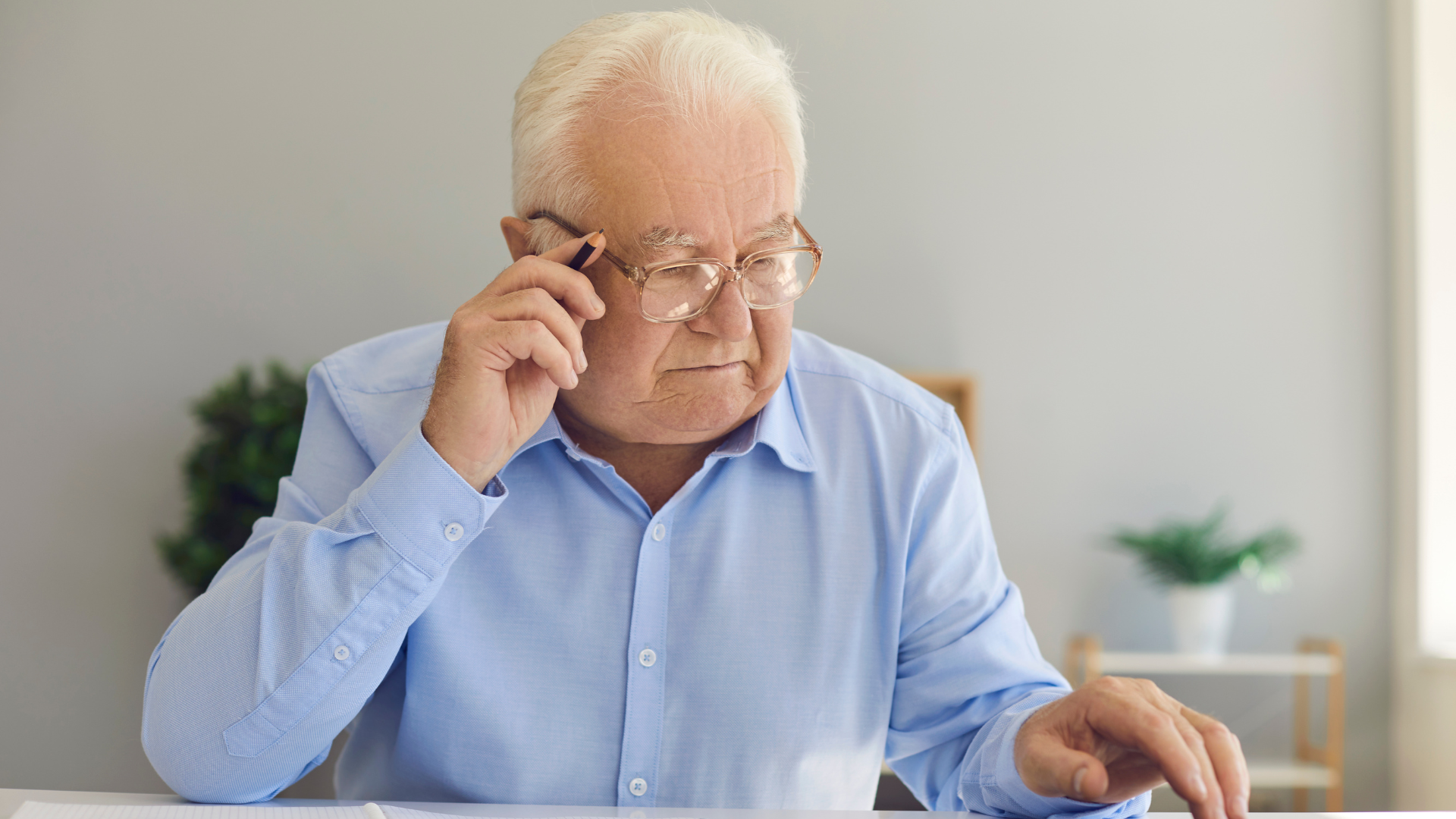 Picture of an older man in a light blue shirt, wearing glasses with a pencil in his right hand. 